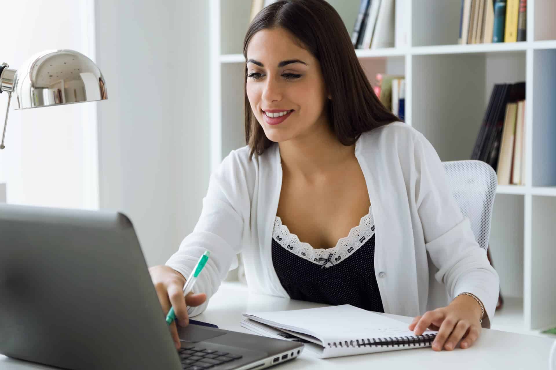 Mujer bonita sentada frente a una laptop, tomando notas en un cuaderno